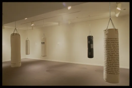 Four white and one black punching bags with text and images printed on them hang distanced from each other from gallery ceiling.