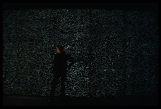 A person in black stands in front of a large wall of bubbles that cast dots of light in a dark space.