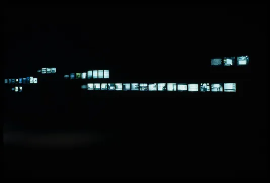 Long shot of a large dark space lit only by scattered rows of active monitors, displaying bluegreen images.