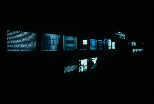 An angled view of rows of monitors displaying bluegreen images of objects, places, and text, casting light in a dark space.