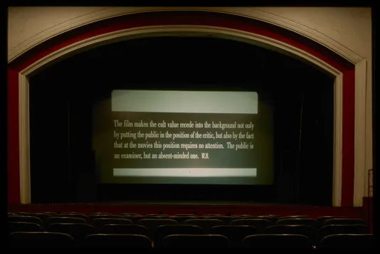 In an unoccupied theater, in the center of the stage, a screen displays a quote on a dark banner, over a blank background.