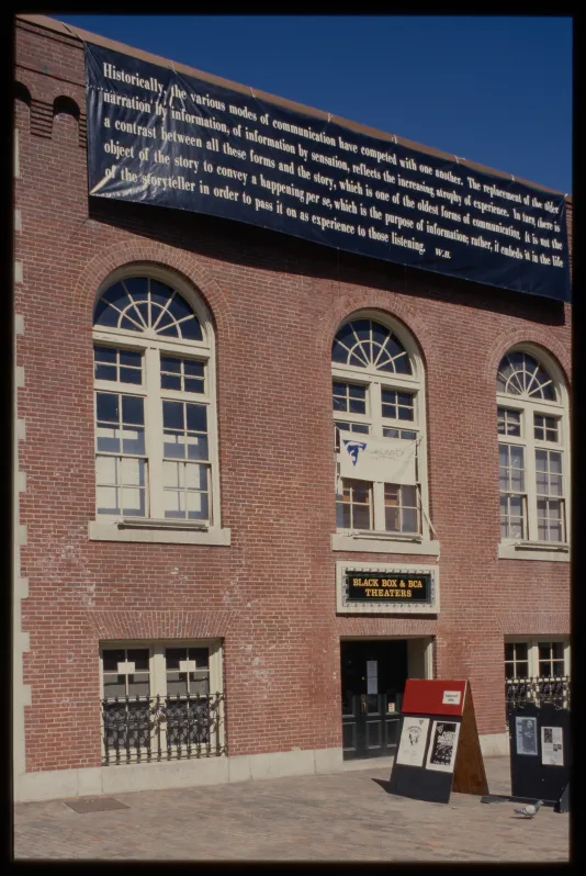 A large black banner with several lines of text hangs above 3 arched windows of a brownstone. A sandwich board sits out front.