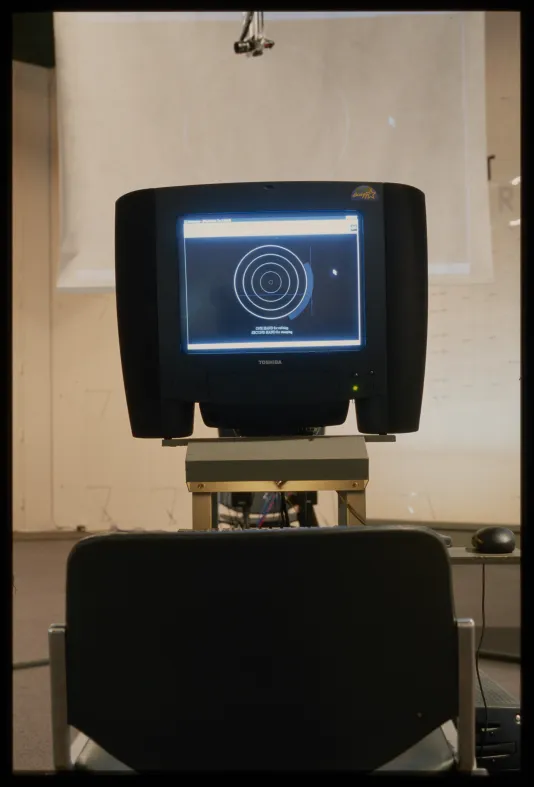 A black monitor displays concentric circles, and is mounted on a metal stand. A film screen is suspended in the background.