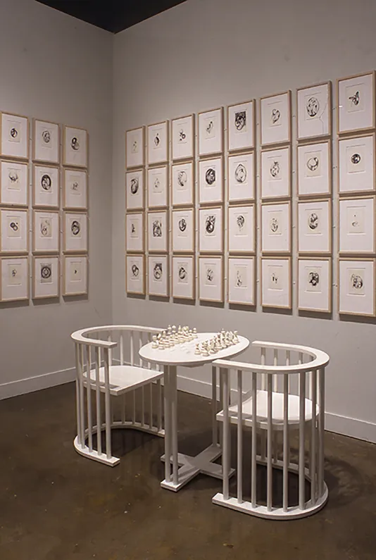 Installation of two white armchairs and a monochromatic chess table in front of a white wall, lined with framed prints 