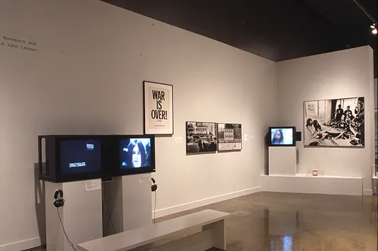 Corner of a white gallery space that includes 3 box videos monitors and and various framed works on the walls