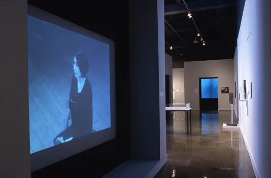 On the right, a video projection of Yoko Ono covers one wall of a walkway that leads into another room with additional artwork