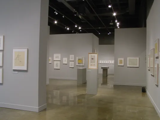 Framed drawings, of various sizes and orientations, hung along multiple light gray walls and placed atop gray pedestals