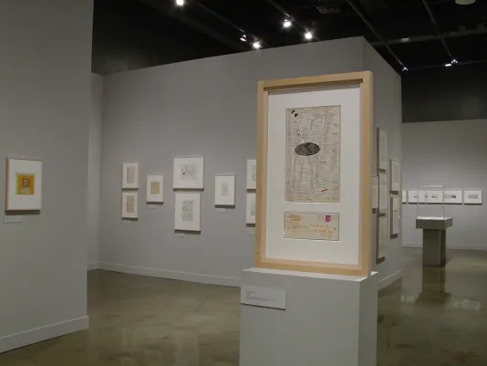 A wooden frame standing atop a gray pedestal. In the background, framed drawings hang on the walls