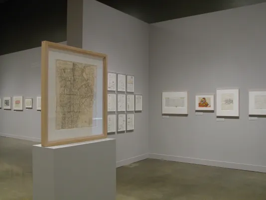 On a pedestal, a wooden frame houses a drawing on yellow paper. Gallery walls are filled with additional framed drawings