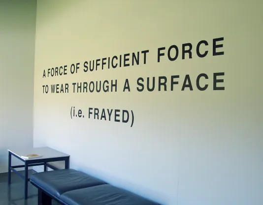 Above a cushioned bench, bold, black wall text reads “A Force of Sufficient Force to Wear Through A Surface (i.e. Frayed)”