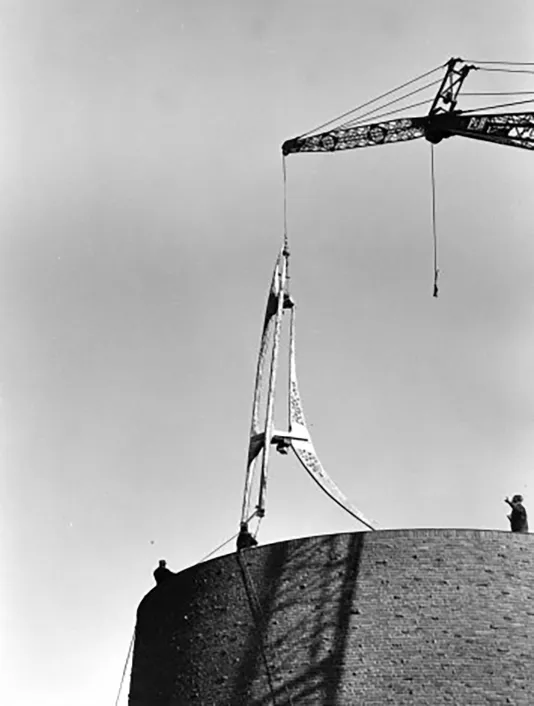 An image depicts the installation of a sculpture atop the MIT Bell Tower. A crane lowers it while onlookers help its placement.