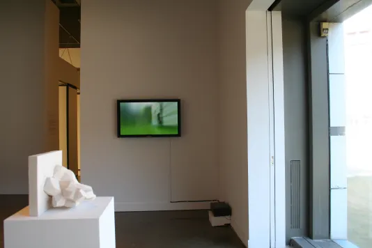 Installation view of white sculpture in front of a monitor against a white wall.