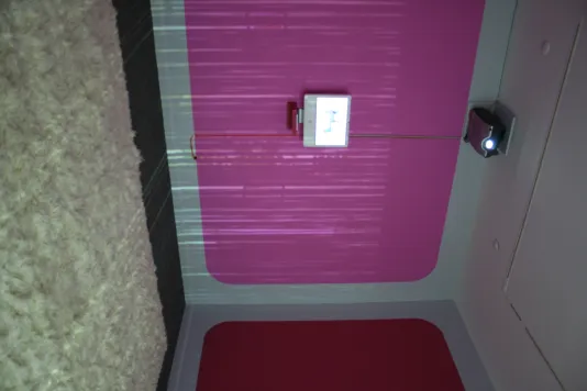 2007 Mac computer monitor affixed to a wall with a pink rectangle with a plush white rug on the ground. 