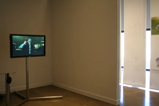 A video monitor screen sits on a stand. On the screen a still from a video of a man inside a recording studio is captured.