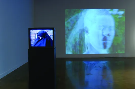 A video monitor w a woman in a headscarf, only part of her face visible and a projection of a boy’s head under a water spray
