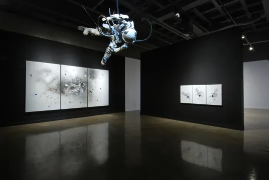 Suspended from a dark ceiling, an astronaut hovers above 2 sets of spot-lit, triptych drawings on perpendicular black walls.