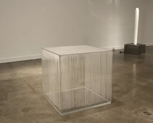 A large cube sculpture made of plexiglass that is covered entirely in condensation on the inside and another sculpture behind.