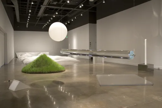 A gallery full of minimal sculptures on the floor and suspended, such as a mound of growing grass and a white balloon.