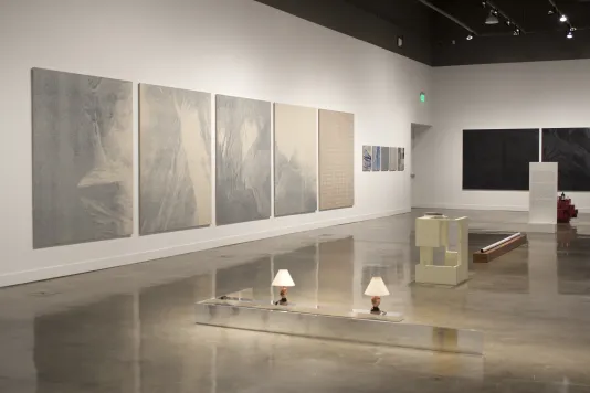 View of gallery with 5 large grey paintings on the left wall and sculptures installed on the floor. 
