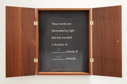 A shallow wood cabinet with chalkboard inset, the words “these words are illuminated by light that has traveled a duration of…”
