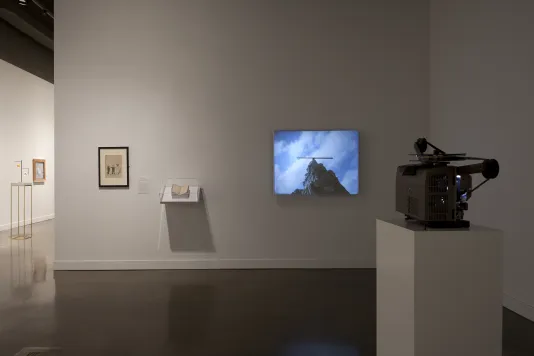 A film projector on a stand, and a screen with image of mountain and sky, a small shelf with an object, a framed picture