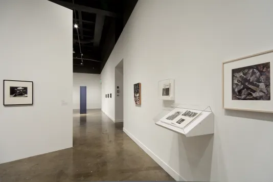 A section of walls with framed pictures, an angled box off the wall with maybe drawings, a black monolith at the far end