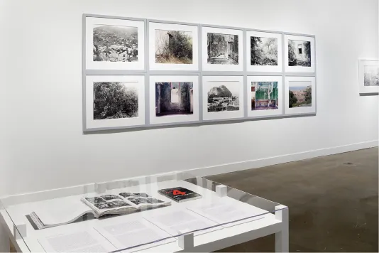 Ten framed artworks line the white wall in a grid of 2 rows. In the foreground, a vitrine table displays books and papers.