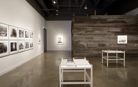 Gallery space with 2 table vitrines and multiple artworks hung on the walls. A wooden wall is built to the right