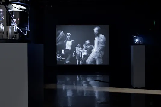 In a large, dark gallery space, a projection shows a man standing still on an otherwise black screen