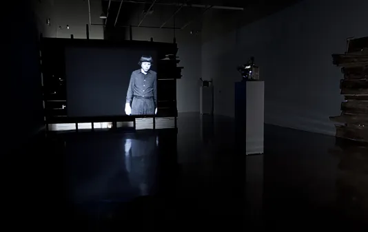 In a large, dark gallery space, a projection shows a man standing still on an otherwise black screen
