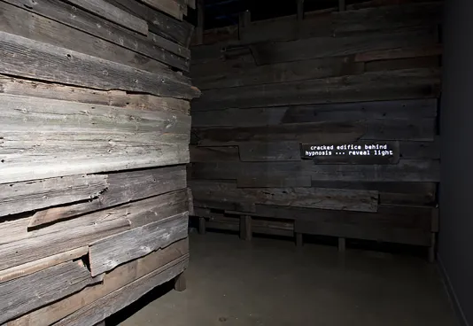 Two walls constructed from gray toned wooden planks meet in a dim corner of the gallery space