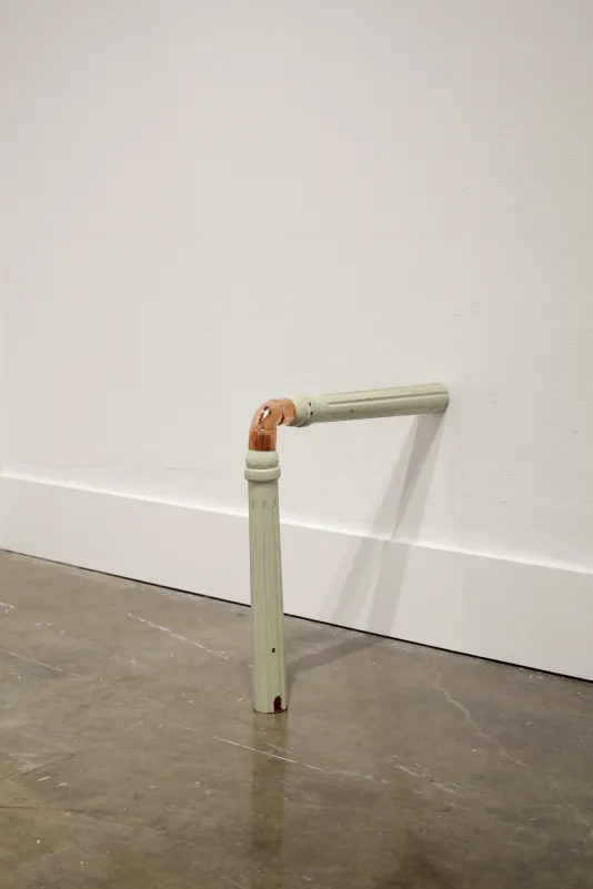 Two painted wooden chair legs, joined by an elbow of copper pipe, connect at the wall and floor at a 90 degree angle.