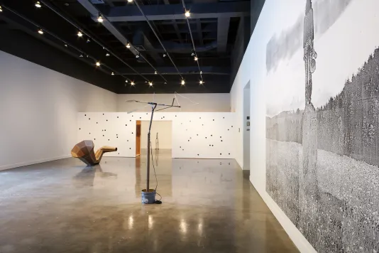 Long view of a gallery displaying a mural in the foreground, 2 sculptures in center, a perforated wall, and a sculpture beyond.