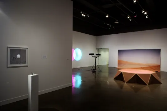 View of a softly lit gallery space, displaying 4 separate artworks of various mediums, using print, cardboard, and light.