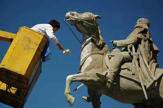Video still of a person in a yellow bucket lift, reaching to paint a statue of a soldier on a horse, against a deep blue sky.
