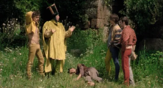 A man in a tall cylindrical hat and a gold tunic talks to 3 young men as they stand over a young man asleep in the grass.