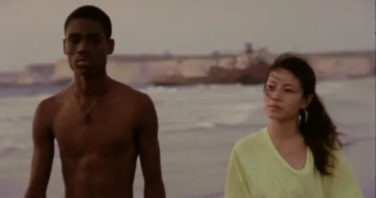 A shirtless dark skinned man and a light skinned woman with long dark hair, walk together as ocean waves break behind them.