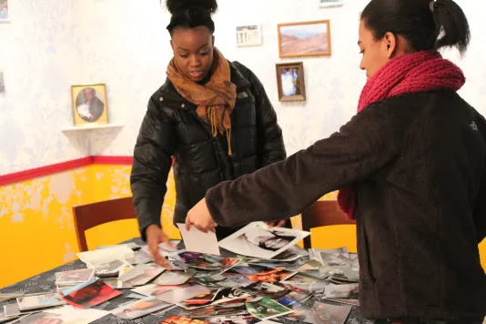 Two young women in coats and scarves stand over a table littered with photos. 1 holds out a photo for the other.