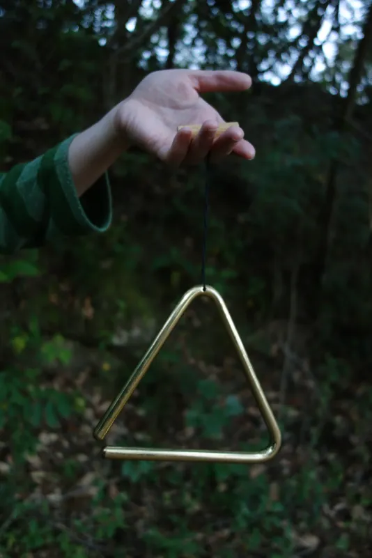 A gold-tone metal triangle dangles from a hand against a blurred background of green vegetation.