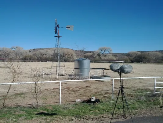 Under a clear blue sky, mics on a stand placed before a white fence, windmill and metal tank in a desert landscape.
