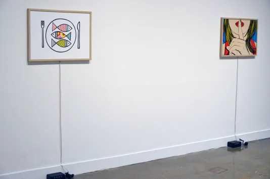 Two colorful prints in wooden frames hang on a white wall, each connected to an exciter and motion detector on the floor.