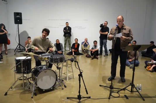 David Grubbs and Eli Keszler perform One and One Less in the gallery before an audience in front of a wall with text in pencil.