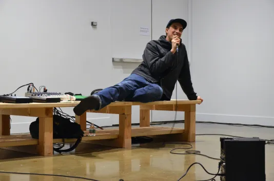 Brendan Fowler performs in the gallery while reclining on a wooden platform. A soundboard, speaker and wires are visible.