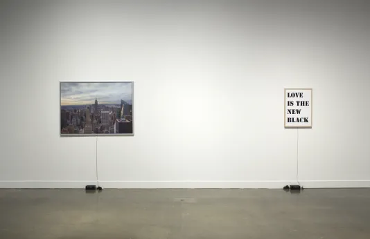 A cityscape hangs to the left of a print reading, “Love is the New Black”. Below each print on the floor is a box and wires.