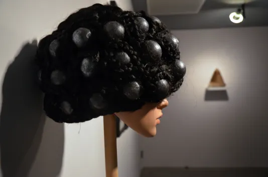 Ping-pong balls painted black embedded in a wig of curly black hair displayed on a female manikin head atop a wood stick