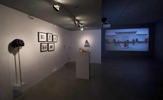Props and drawings displayed in left side of gallery and on the right, a large projection of people walking before a building