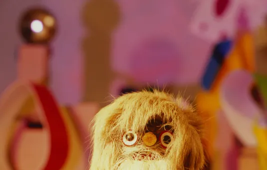 Against a blurred, colorful backdrop, a yellow furry creature with plastic googly eyes peers from the bottom of the frame.