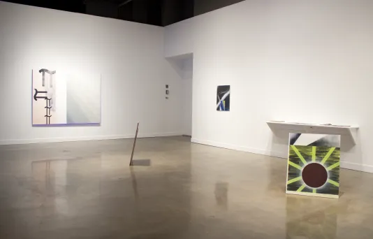 Two paintings are displayed on the wall and 2 on the floor. A wall-mounted shelf with objects upon it is also visible.