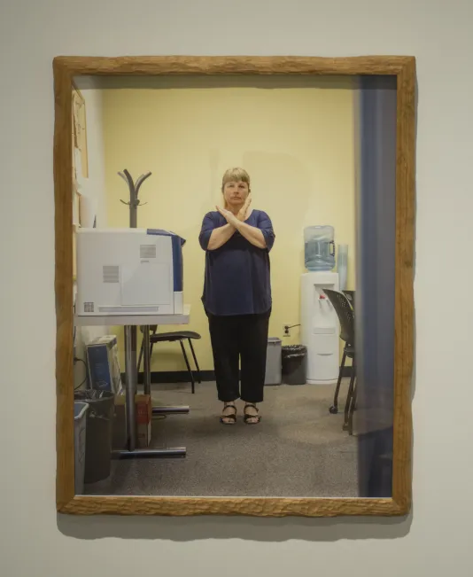 A photo in a brown frame features a woman standing in an office next to a printer and the water cooler with her arms held up in an X shape.