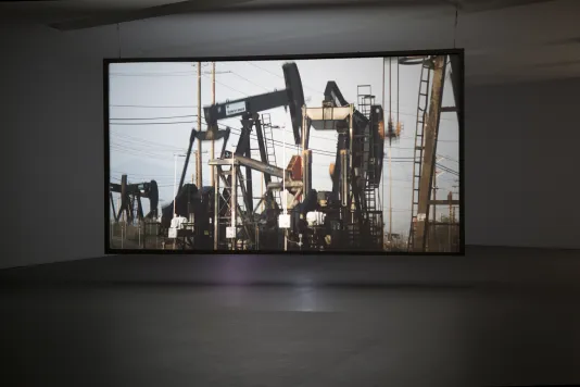 Large oil drilling rigs against a murky blue sky is projected on a large cinematic screen in a dark gallery.